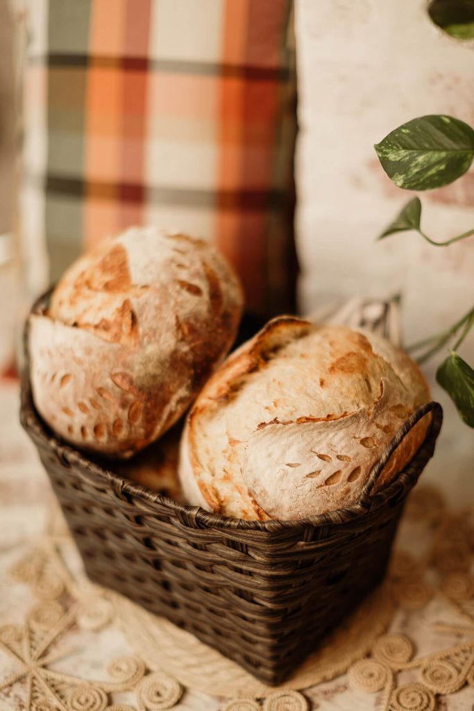 A dark brown woven basket holds two round, crusty sourdough loaves, both featuring golden crusts and decorative wheat or leaf scoring. The basket rests on a crocheted or macrame mat, with a blurred background showing an orange and brown plaid pillow and a green houseplant.